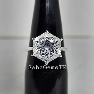 May include: A silver ring with a large, clear, hexagonal-cut gemstone. The gemstone has an intricate, geometric pattern within. The ring is displayed on a black stand. The text "SabaGemsIN" is visible.