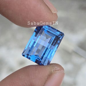 以下が含まれることがあります： 長方形のエメラルドカットの鮮やかな青い宝石。宝石は手で持っており、その透明度と輝きが際立っています。