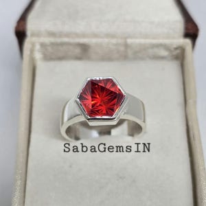 May include: A silver ring with a red hexagonal gemstone. The ring is in a white box with the text "SabaGems IN".