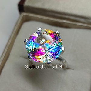 May include: A silver ring with a large, round, multi-colored gemstone. The gemstone has a rainbow of colors, including red, orange, yellow, green, blue, and purple. The ring is in a small, white box.