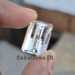 Peut inclure: Une pierre précieuse rectangulaire transparente avec de multiples facettes, tenue entre les doigts. La gemme est transparente, reflétant la lumière et créant un effet scintillant. Le texte "SabaGems IN" est visible en bas. Cette pierre est probablement pour la joaillerie.