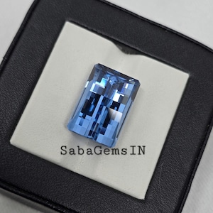 May include: A light blue gemstone with a rectangular cut, displayed on a black and white background. The gemstone has a faceted design and is set in a clear setting. The text "SabaGemsIN" is visible in the bottom centre of the image.