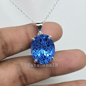 May include: A silver necklace with a large, oval, faceted blue gemstone pendant. The pendant is held by four silver prongs and has intricate internal patterns. The chain is a delicate silver link. The text "SabaGems IN" is visible.