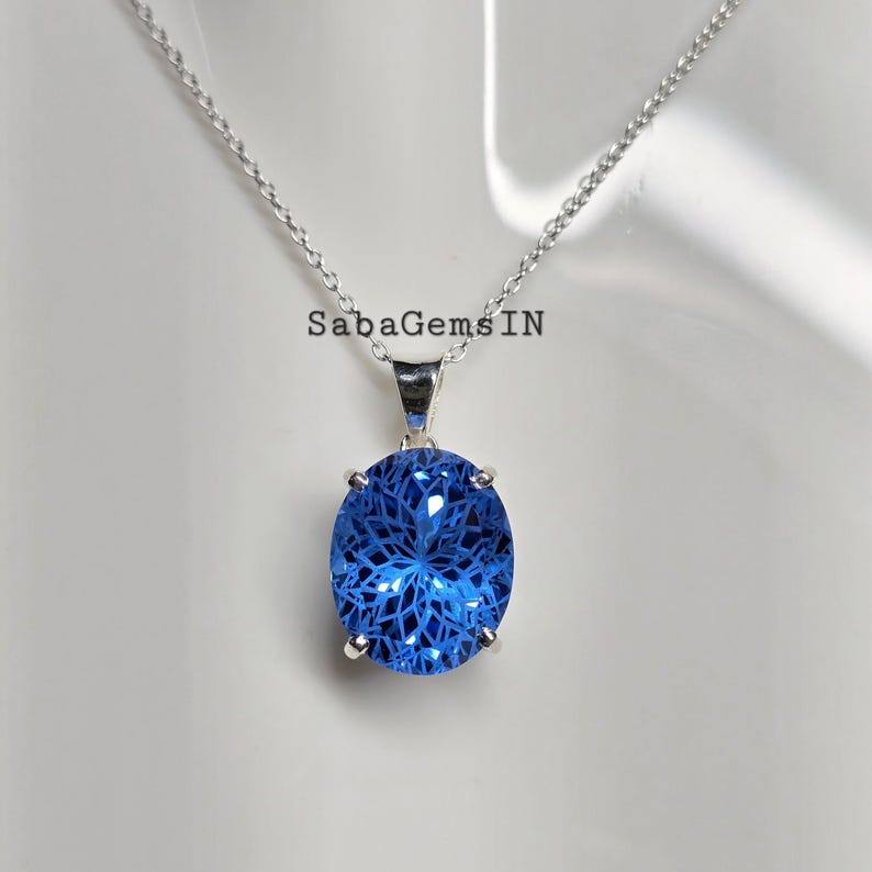 May include: A silver necklace with a large, oval-shaped blue pendant. The pendant has an intricate, web-like design within the blue gemstone. The necklace is displayed against a white background. The text "SabaGems IN" is visible.