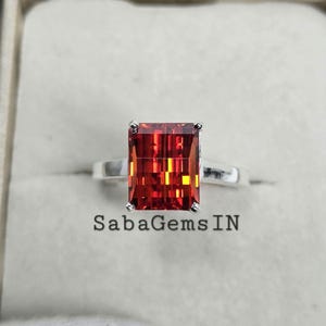 May include: A silver ring featuring a large, rectangular, red gemstone. The gemstone has a faceted cut and a deep red colour with orange and yellow hues. The ring is set in a simple band and has a polished finish. The ring is displayed on a white background.  SabaGemsIN is printed below the ring.