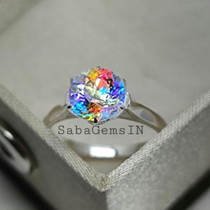 May include: A silver ring with a large, round, iridescent gemstone. The gemstone displays a rainbow of colors, including red, orange, yellow, green, blue, and purple. The ring is set in a simple, elegant band. The text "SabaGemsIN" is visible on the ring.
