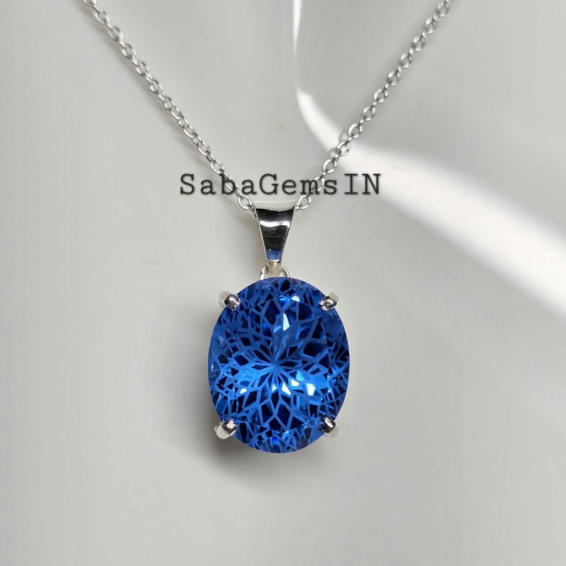 May include: A silver necklace with a large, oval, faceted blue gemstone pendant. The pendant is suspended from a delicate chain. The text "SabaGems IN" is visible above the pendant.