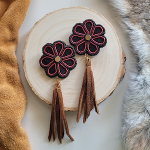 Può includere: Un paio di orecchini a forma di fiore nero con perline e dettagli rossi e nappe in pelle scamosciata marrone.