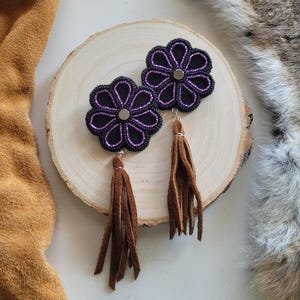 Può includere: Un paio di orecchini a forma di fiore con accenti viola e neri e nappe in pelle scamosciata marrone.