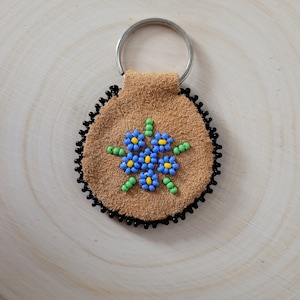 May include: A tan suede leather keyring with a silver ring. It features a beaded floral design with blue, yellow, and green beads. The edge is trimmed with black beads.