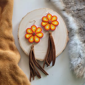 Può includere: Un paio di orecchini a forma di fiore perlato con nappe in pelle scamosciata marrone. I fiori sono gialli, rossi e bianchi con un centro dorato.