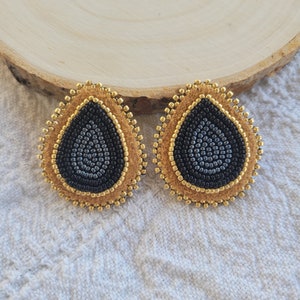May include: A pair of teardrop-shaped earrings with a black beaded center surrounded by a gold beaded border and a tan suede backing.