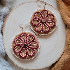 Può includere: Un paio di orecchini fatti a mano con un design floreale. Gli orecchini hanno una base in pelle scamosciata marrone, adornata con perline rosse e rosa a forma di fiore. Perline bianche delineano i bordi, con dettagli dorati e ganci dorati.