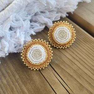 Puede incluir: Un par de pendientes redondos de color dorado con un centro de cuentas blancas y beige. Los pendientes están sobre una superficie de madera.