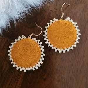 Può includere: Un paio di orecchini in pelle scamosciata color senape con dettagli di perline bianche. Gli orecchini sono rotondi e hanno un design semplice.