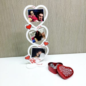 Può includere: Una cornice bianca a forma di cuore con tre aperture, ciascuna contenente una fotografia. Cuori rossi e la parola "LOVE" decorano la cornice. Una scatola di cioccolatini rossa a forma di cuore con la scritta "Happy Valentine's Day" è vicina.