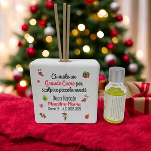 Può includere: Un diffusore quadrato bianco con bastoncini di legno, una piccola bottiglia di liquido giallo e decorazioni natalizie. Il diffusore presenta il testo "Ci vuole un Grande Cuore per scolpire piccole menti Buon Natale Maestra Maria Classe 1A - A.S. 2023-2024."