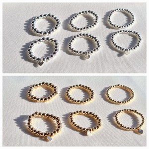 Könnte beinhalten: Sechs silberne und sechs goldene Perlenringe mit einem kleinen Herzanhänger an jedem Ring. Die Ringe sind in zwei Reihen zu je drei angeordnet.
