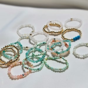 Peut inclure: Une collection de bagues en perles de différentes couleurs, notamment blanc, turquoise et rose. Les bagues sont composées de petites perles rondes et sont empilées les unes sur les autres.