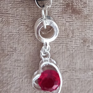 May include: A silver heart-shaped pendant with a red gem inside, attached to a black elastic hair tie.