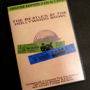 May include: A DVD case with a yellow background and a picture of The Beatles. The case says "Deluxe Edition 3 CD & 1 DVD The Beatles at the Hollywood Bowl". It also has two ticket stubs for The Beatles at the Hollywood Bowl, one for August 23rd and one for August 29th. The text on the case says "Contains all 3 concerts plus the original 1977 album release & a DVD with concert footage and more!"