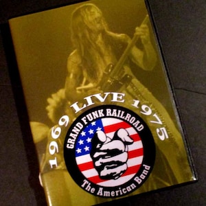 May include: A VHS tape with a yellow background and a black circle in the center. The circle has the text "Grand Funk Railroad" and "The American Band" in white. The circle also has a red, white, and blue American flag with a black fist in the center. The text "Live 1969-1975" is in white and surrounds the circle.