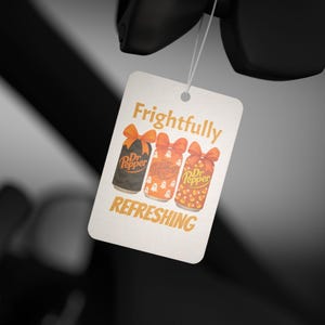 May include: A white tag with the words "Frightfully Refreshing" and three Dr. Pepper cans with orange bows. The cans are decorated with Halloween themes: a black can, an orange can with ghosts, and an orange can with candy corn.