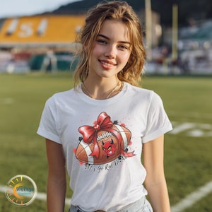 May include: White t-shirt featuring a football design with a red bow, a devil face, and the text "Let's go Red Devils." The football is brown with white stripes. The design has red and black accents.
