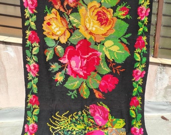 May include: A black wool rug with a floral design featuring red, yellow, and pink roses. The rug has a green border with small pink roses.