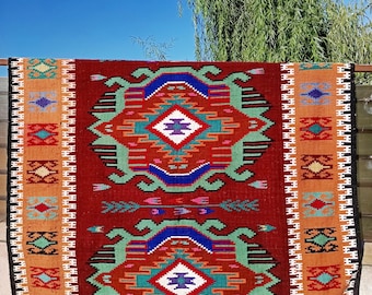 May include: A rectangular woven rug featuring a deep red central field with two large, symmetrical geometric diamond motifs in blue, green, white, and purple. The rug is framed by wide orange borders decorated with repeating colorful geometric patterns.