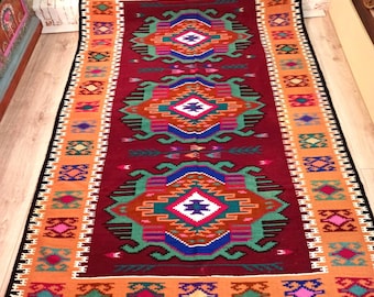 May include: A colourful, hand-woven rug with a geometric pattern in shades of red, orange, green, blue, and white. The rug has a fringed border and is made of wool.