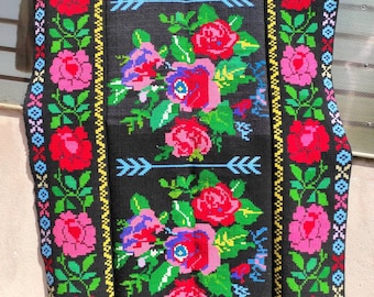 May include: Black textile with a floral design featuring red, pink, and purple roses, green leaves, and blue arrows. The edges are adorned with a border of colourful geometric patterns and floral motifs.