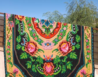 May include: A colourful, rectangular rug with a floral pattern. The rug has a black border, a peach-coloured background, and vibrant flowers in pink, red, green, and blue. The rug is hanging outdoors against a blue sky.
