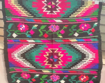 May include: A vibrant, rectangular woven rug with a black background and colorful geometric patterns. The design features pink, green, yellow, and white floral and geometric shapes. The rug is displayed outdoors.