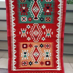 Könnte beinhalten: Ein rechteckiger, gewebter Wandbehang mit rotem Hintergrund und geometrischen Mustern in Grün, Orange, Schwarz und Weiß. Das Design umfasst Diamanten- und Sternformen mit einem gefransten unteren Rand. Der Behang ist etwa 76 cm lang.