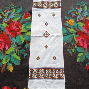 Könnte beinhalten: Weißes rechteckiges Textil mit gestickten geometrischen Mustern in Rot, Grün, Gelb und Schwarz. Das Textil hat Fransen an den Kanten und wird auf einem floral gemusterten Hintergrund präsentiert. Die Stickerei zeigt Diamant- und Kreuzmotive.