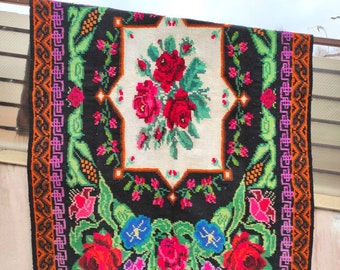 May include: A black wool rug with a white centre featuring a floral design of red, pink, and white roses. The rug has a colourful border with green, orange, and purple accents.
