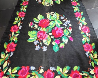 May include: A black wool rug with a floral pattern featuring pink, red, green, and yellow roses. The rug has a fringe border.