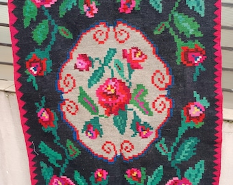 May include: A rectangular, dark gray rug with a central floral design. The rug features a pink border with a zigzag pattern. The floral design includes red and pink roses, green leaves, and a gray background.