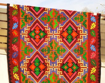 May include: A colourful handwoven rug with a geometric pattern in shades of red, green, blue, yellow, and pink. The rug has a fringed border.