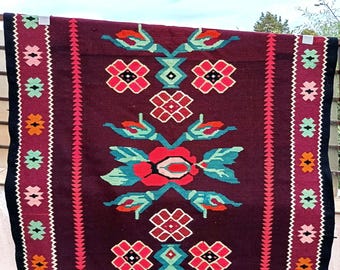 May include: A traditional handwoven rug with a burgundy background and a colourful floral pattern. The rug has a fringe border and is made of wool.