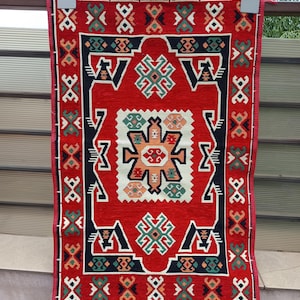 Könnte beinhalten: Ein roter rechteckiger Teppich mit einem detaillierten geometrischen Muster. Der Teppich hat ein zentrales Design mit weißem Rand, umgeben von schwarzen, grünen und orangefarbenen Akzenten. Die Kanten sind mit einem roten Fransen versehen.