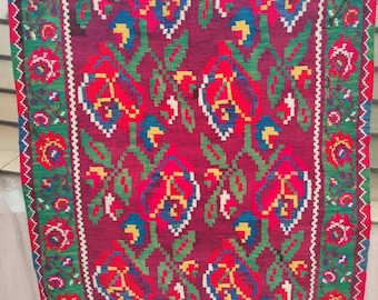 May include: A traditional red, green, blue, and white patterned rug with a floral design. The rug has a fringe border.