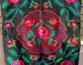 May include: A dark textile with a vibrant floral design. The central motif is a circular pattern in red and blue, surrounded by pink and red roses with green leaves. The background is black.