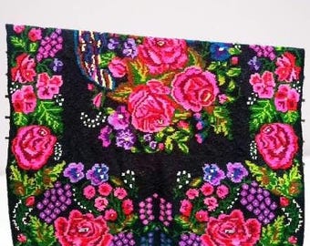 May include: A black fabric rug with a floral design featuring pink, red, and white roses, green leaves, and purple grapes. The rug has a fringe border.