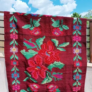 Könnte beinhalten: Ein burgunderfarbener, rechteckiger Teppich mit floralem Design. Der Teppich zeigt große rote Blumen mit grünen Blättern in der Mitte und einen Rand aus kleineren Blumen in Rosa, Blau und Gelb. Der Teppich hängt draußen vor blauem Himmel.