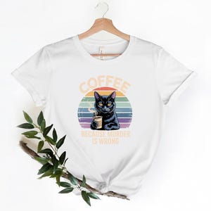 May include: White t-shirt featuring a black cat holding a coffee cup, set against a colorful sunset design. The text "COFFEE BECAUSE MURDER IS WRONG" is printed above and below the cat illustration. The shirt is hanging on a wooden hanger.