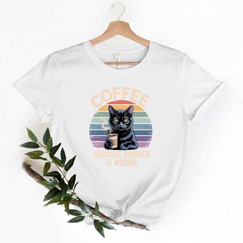 May include: White t-shirt featuring a black cat holding a coffee cup, set against a colorful sunset design. The text "COFFEE BECAUSE MURDER IS WRONG" is printed above and below the cat illustration. The shirt is hanging on a wooden hanger.