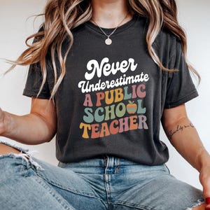 Peut inclure: T-shirt gris foncé avec l'inscription "Never Underestimate A PUBLIC SCHOOL TEACHER" en blanc, rose, orange, vert et violet. Le t-shirt a un style rétro et vintage.