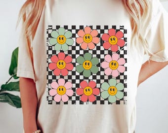 May include: A cream-coloured t-shirt with a vibrant floral design. The design features nine smiling flowers in pink, green, and orange, set against a black and white chequered background. The flowers have yellow smiley faces.